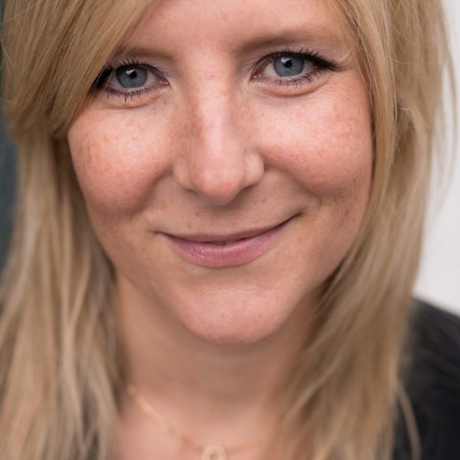 Frau mit blauer gestreifter Bluse lächelt, Hände gefaltet, in einem hellen Büro.