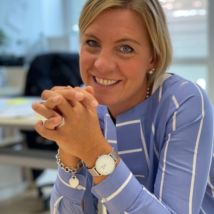 Frau mit blauer gestreifter Bluse lächelt, Hände gefaltet, in einem hellen Büro.